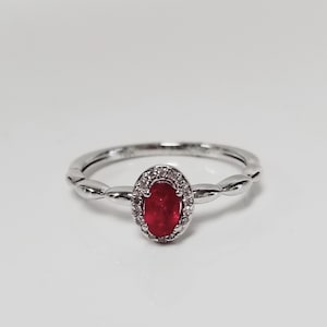 May include: A silver ring featuring a red oval gemstone set within a halo of smaller white gemstones.