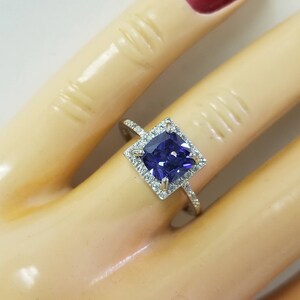 Op de afbeelding: Een zilveren ring met een vierkante paarse edelsteen omringd door kleinere witte edelstenen.