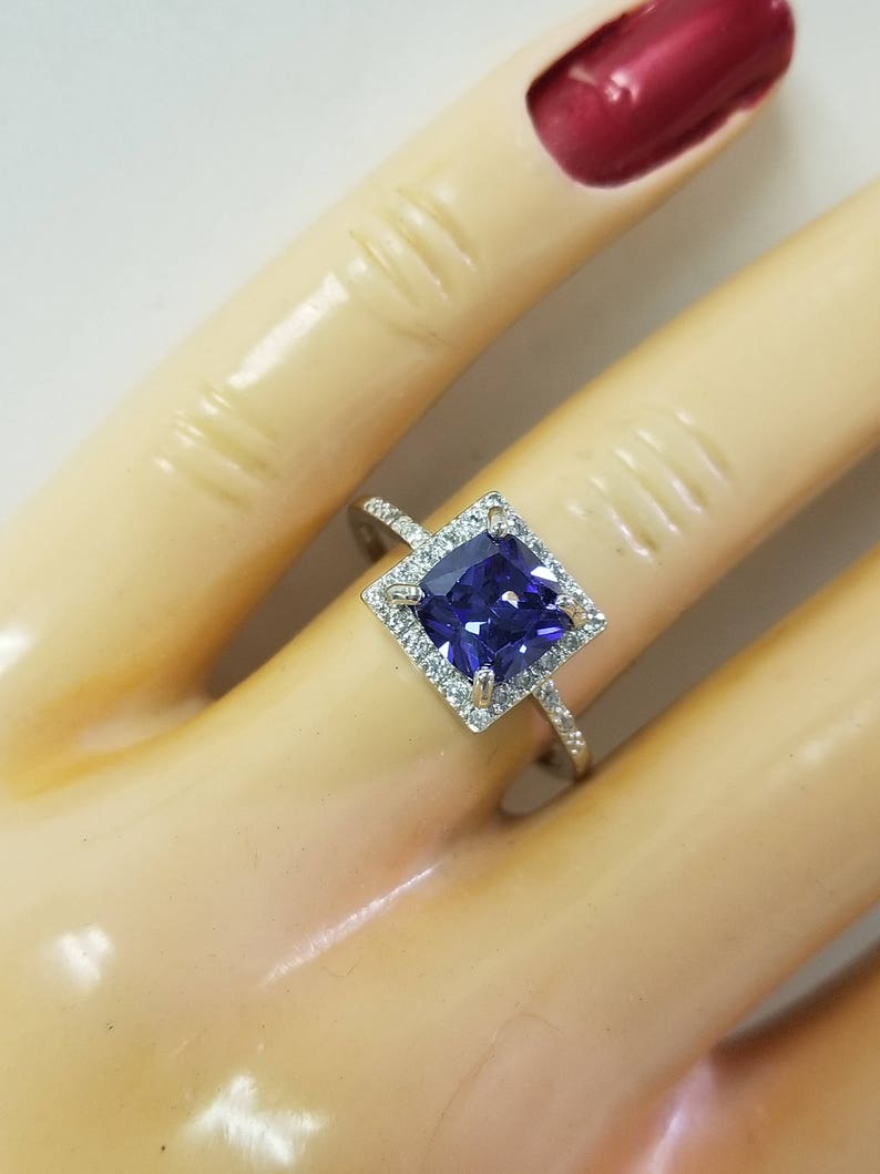 Op de afbeelding: Een zilveren ring met een vierkante paarse edelsteen omringd door kleinere witte edelstenen.