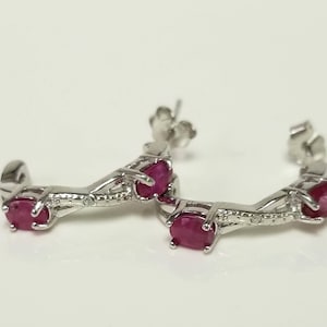 May include: A pair of silver-toned earrings featuring a curved, open-hoop design. Each earring is set with two oval-shaped, deep red rubies held in prong settings, connected by a textured, metallic bridge. The earrings include standard post-back closures.