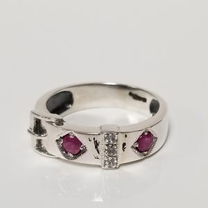May include: A silver ring with a buckle design. The ring features two red gemstones in diamond-shaped settings and a central section with three smaller clear stones. The ring is set against a white background.