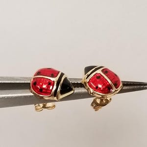 May include: A pair of ladybug stud earrings. Each earring features a red enamel body with black spots and a black head, outlined in gold. The earrings are held by a silver tool against a white background.