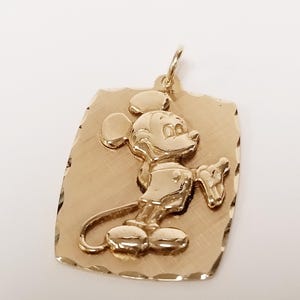 K&ouml;nnte beinhalten: Goldfarbener Anh&auml;nger mit erhabenem Mickey Mouse-Motiv. Der Anh&auml;nger hat eine rechteckige Form mit strukturierter Oberfl&auml;che und einer &Ouml;se f&uuml;r eine Kette. Die Zeichentrickfigur ist in einer klassischen Pose dargestellt.
