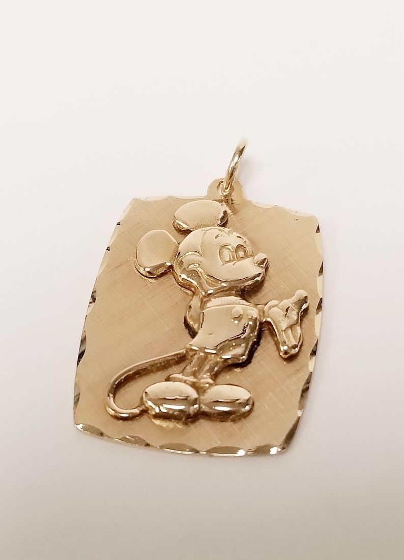 K&ouml;nnte beinhalten: Goldfarbener Anh&auml;nger mit erhabenem Mickey Mouse-Motiv. Der Anh&auml;nger hat eine rechteckige Form mit strukturierter Oberfl&auml;che und einer &Ouml;se f&uuml;r eine Kette. Die Zeichentrickfigur ist in einer klassischen Pose dargestellt.