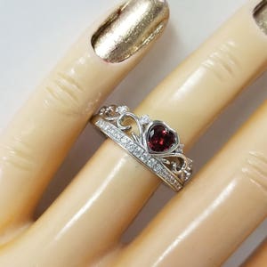 May include: A silver ring featuring a heart-shaped red gemstone surrounded by smaller clear gemstones. The ring is designed in a crown shape.