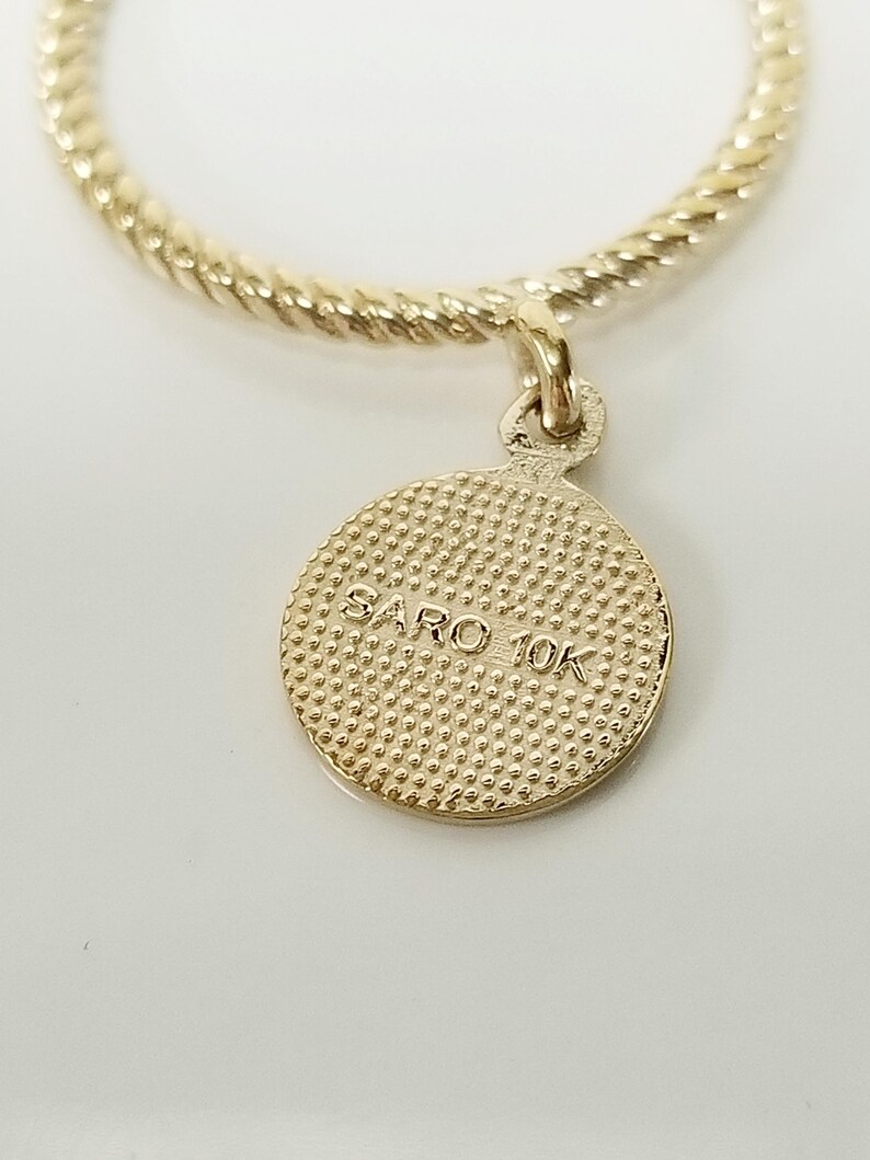 May include: A gold-colored necklace with a twisted chain and a round pendant. The pendant has a textured surface and the text "SARO 10K" inscribed on it.