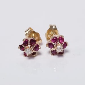 May include: A pair of gold-toned stud earrings. Each earring features a flower design with six deep red gemstones surrounding a central clear gemstone. The earrings are set against a white background.