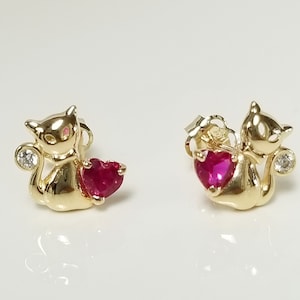 May include: A pair of gold-colored cat-shaped stud earrings. Each cat holds a heart-shaped, red gemstone and has a small, clear gemstone near its tail. The earrings are displayed on a white surface.