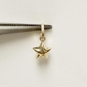 Könnte beinhalten: Ein goldfarbener Stern-Anhänger mit einer Öse oben zum Anbringen an einer Halskette oder einem Armband.