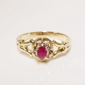May include: A gold ring with an oval, deep red gemstone at its centre, encircled by small, clear stones. The ring's band has a decorative design with a split shank. The ring is set against a white background.