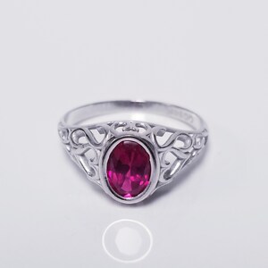May include: A silver ring featuring a central oval-cut vibrant pink gemstone. The band is crafted with an intricate openwork filigree design that extends from the setting, providing a delicate and detailed appearance to the metalwork surrounding the stone.
