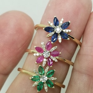 May include: Three gold rings with flower-shaped settings. The top ring has a blue sapphire centre stone surrounded by smaller diamonds. The middle ring has a pink ruby centre stone surrounded by smaller diamonds. The bottom ring has a green emerald centre stone surrounded by smaller diamonds.