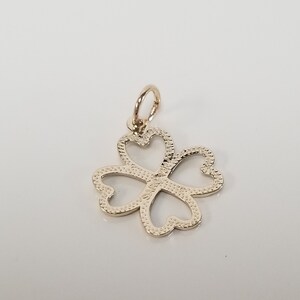 May include: A gold-colored four-leaf clover charm pendant with a textured surface. The charm has a loop at the top for attaching to a necklace or bracelet.