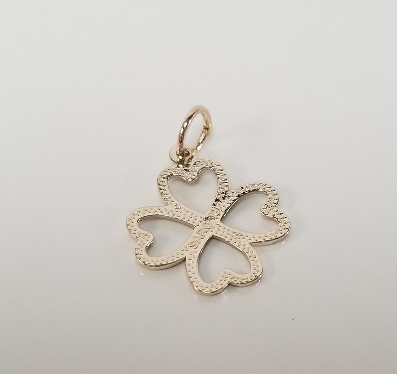 May include: A gold-colored four-leaf clover charm pendant with a textured surface. The charm has a loop at the top for attaching to a necklace or bracelet.