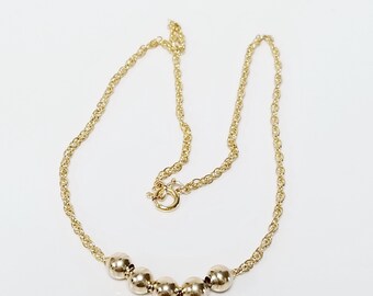 May include: A gold-colored necklace with a delicate chain and five evenly spaced, round beads. The necklace has a simple clasp and is displayed against a white background. The jewelry is likely made of gold or a gold-toned metal.