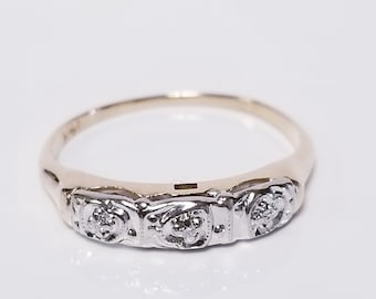 May include: A gold-toned ring with three small, clear gemstones set in a decorative, vintage-style band. The ring is centered on a white surface, showcasing its intricate design and reflective qualities.