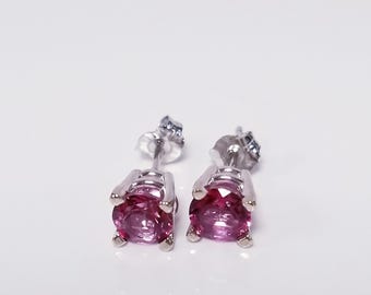 May include: A pair of stud earrings featuring round, deep pink gemstones set in silver-colored metal. The earrings have a classic, minimalist design, perfect for everyday wear or special occasions.