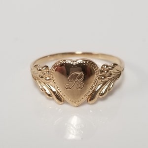 Op de afbeelding: Een gouden ring in de vorm van een hart met een delicaat bloemmotief aan de zijkanten. Het hart heeft een gegraveerde letter "R" in het midden.
