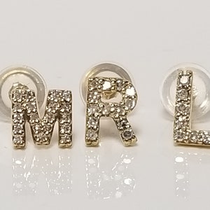 May include: Three gold-colored stud earrings spelling out the letters M, R, and L. Each letter is encrusted with small, sparkling gemstones. The earrings are displayed on a white surface.