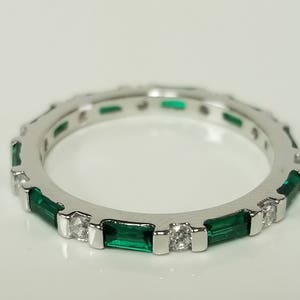 Op de afbeelding: Een zilveren ring met afwisselend smaragdgroene edelstenen en kleine heldere stenen. De ring is geplaatst tegen een witte achtergrond, die het ontwerp en de glans benadrukt. De ring is een sieraad.
