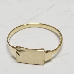 May include: A gold ring featuring a rectangular, flat-topped plate as the centerpiece. The plate has a brushed, matte finish and a small, engraved leaf-like detail on the left side. The band is thin, smooth, and circular, connecting to the plate.