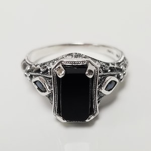 May include: A silver ring featuring a central rectangular black stone held by four prongs. The band and setting are crafted with intricate metalwork, including small decorative side stones and detailed filigree patterns along the shank.