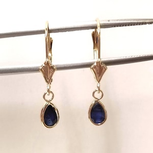 May include: A pair of gold leverback earrings featuring a diamond-shaped metal top piece connected to a dangling teardrop-shaped blue sapphire gemstone set in a gold bezel. The earrings are suspended from a metal tool for display purposes.