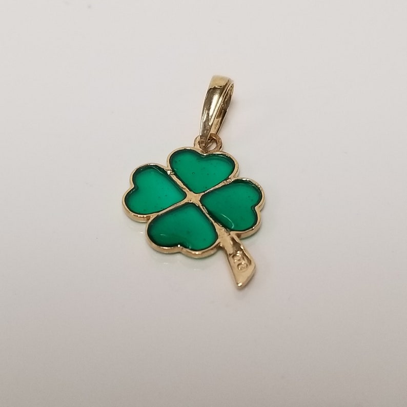 May include: A gold-colored pendant featuring a four-leaf clover design. The clover's leaves are a vibrant green, outlined in gold. The pendant has a small loop for a chain and a textured stem.