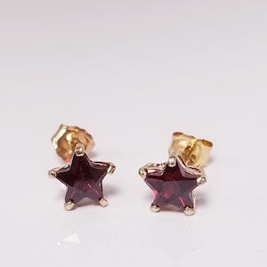 May include: A pair of gold-coloured star-shaped stud earrings. Each earring features a deep red gemstone set within the star's frame. The earrings are displayed on a white surface, showcasing their design and colour.