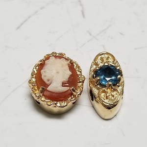 Könnte beinhalten: Zwei goldfarbene Anhänger. Einer zeigt eine Kamee mit dem Profil einer Frau in einem rotbraunen Rahmen. Der andere Anhänger hat einen blauen Edelstein in einem verzierten Goldrahmen. Schmuck.