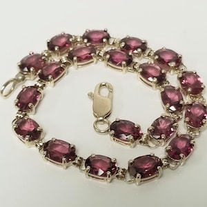 May include: A silver-toned bracelet featuring a series of oval-shaped, deep magenta-colored faceted stones set in metal prongs. The bracelet is arranged in a circular shape and includes a silver-toned lobster claw clasp mechanism for closure.