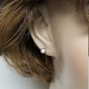 May include: A close-up of a small, round, clear gemstone earring with a gold setting. The earring is worn in the left ear of a mannequin.