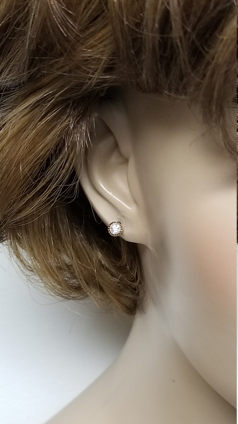 May include: A close-up of a small, round, clear gemstone earring with a gold setting. The earring is worn in the left ear of a mannequin.