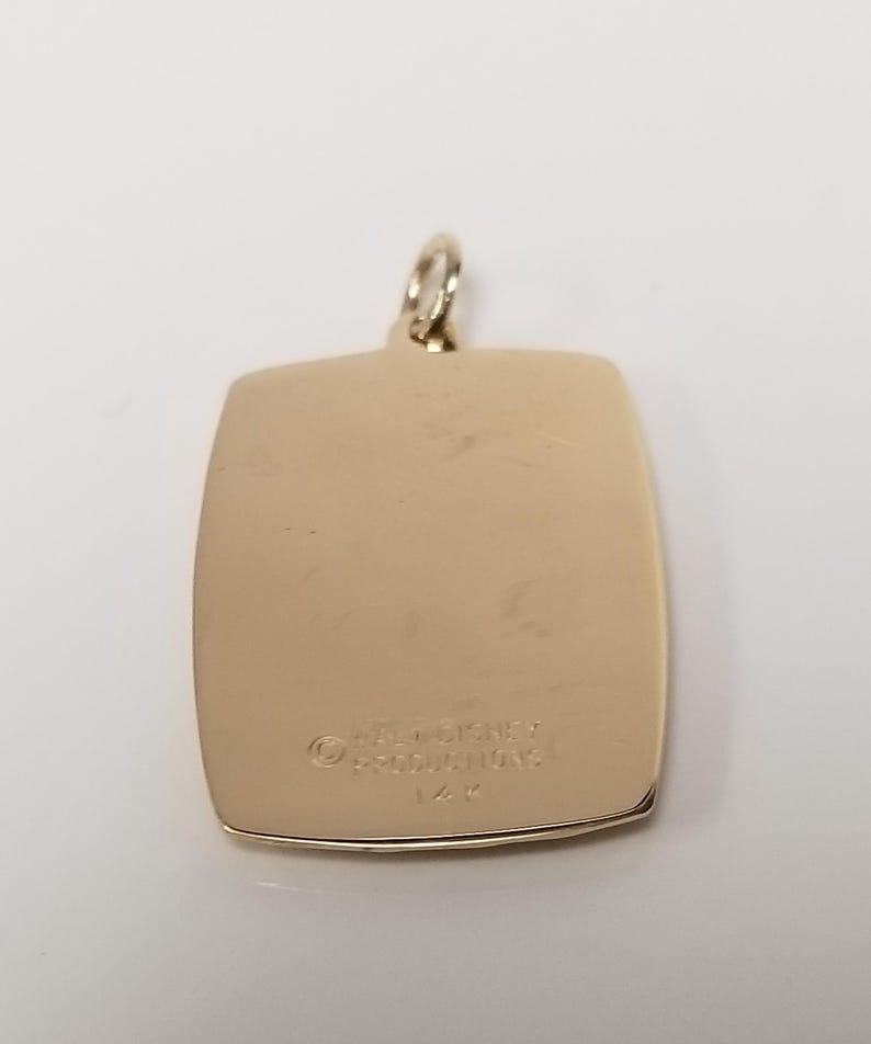 K&ouml;nnte beinhalten: Ein goldfarbener rechteckiger Anh&auml;nger mit abgerundeten Ecken und einer kleinen Schlaufe oben. Der Anh&auml;nger tr&auml;gt den Text "WALT DISNEY PRODUCTIONS" und "14K" auf der R&uuml;ckseite eingraviert.
