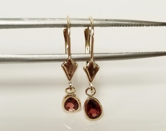 May include: A pair of gold-colored drop earrings with teardrop-shaped, reddish-pink gemstones. The earrings are held by a silver-colored tool against a white background. The earrings have a geometric design.