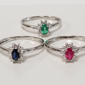 May include: Three silver rings with a white gold setting. Each ring features a different gemstone: a green emerald, a blue sapphire, and a red ruby. Each gemstone is surrounded by a halo of smaller diamonds.
