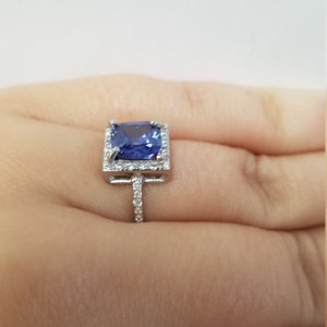 Op de afbeelding: Een zilveren ring met een grote, vierkante blauwe edelsteen omringd door kleinere, heldere edelstenen.