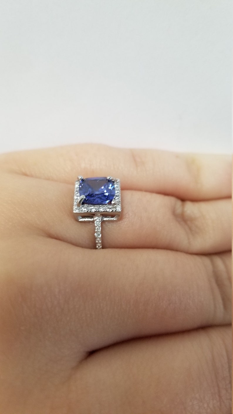 Op de afbeelding: Een zilveren ring met een grote, vierkante blauwe edelsteen omringd door kleinere, heldere edelstenen.