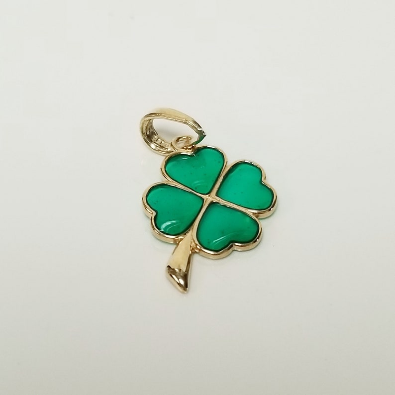 May include: A gold-colored pendant featuring a four-leaf clover design. The clover's four heart-shaped leaves are a vibrant green, outlined in gold. The pendant has a loop for a chain and a stem detail.