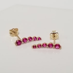 May include: A pair of gold earrings with a row of pink gemstones. The earrings are designed in a curved shape.