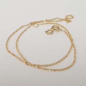 May include: A delicate gold-coloured necklace with a twisted rope chain design. The necklace features a small clasp and a circular attachment. The jewellery is displayed on a white surface, highlighting its intricate details.