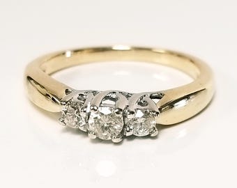 May include: A gold and white gold ring with three round diamonds set in a row. The ring has a polished finish and is set against a white background. The ring is a piece of jewelry.