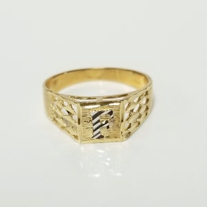 May include: A gold ring with a square-shaped setting featuring the letter "F" in a white metal. The ring has a delicate, intricate design.