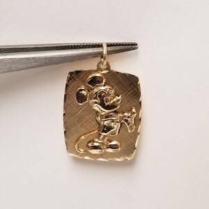 K&ouml;nnte beinhalten: Goldfarbener Anh&auml;nger in rechteckiger Form mit strukturierter Oberfl&auml;che. Der Anh&auml;nger zeigt ein detailliertes Relief von Mickey Mouse, einer klassischen Zeichentrickfigur. Der Anh&auml;nger wird von einer Pinzette gehalten.