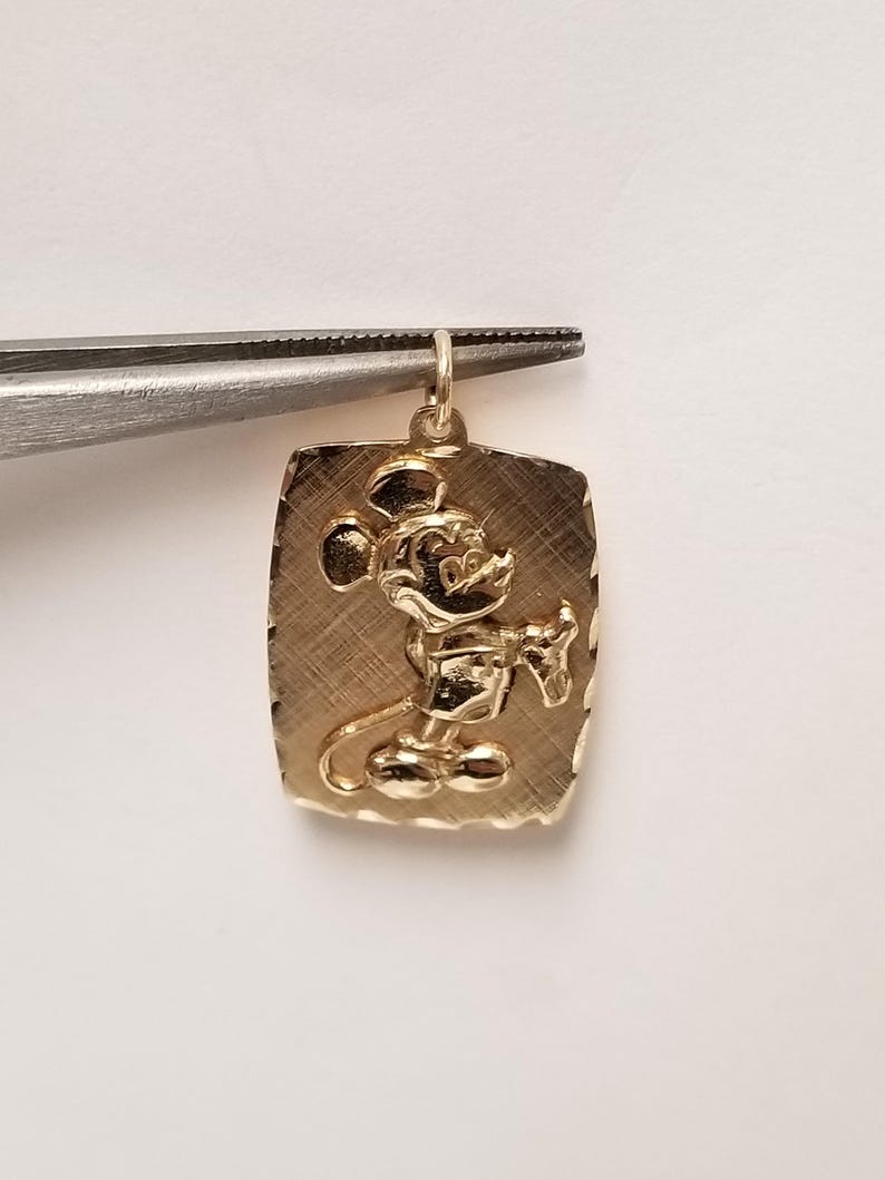 K&ouml;nnte beinhalten: Goldfarbener Anh&auml;nger in rechteckiger Form mit strukturierter Oberfl&auml;che. Der Anh&auml;nger zeigt ein detailliertes Relief von Mickey Mouse, einer klassischen Zeichentrickfigur. Der Anh&auml;nger wird von einer Pinzette gehalten.