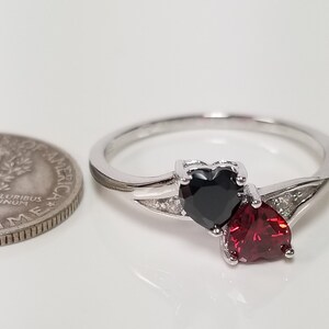 May include: A silver ring with two heart-shaped gemstones. The larger gemstone is red, and the smaller gemstone is black. The ring is set with small clear stones.
