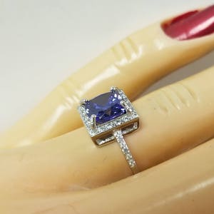 Op de afbeelding: Een zilveren ring met een grote, vierkante, paarse edelsteen omringd door kleinere, heldere edelstenen.