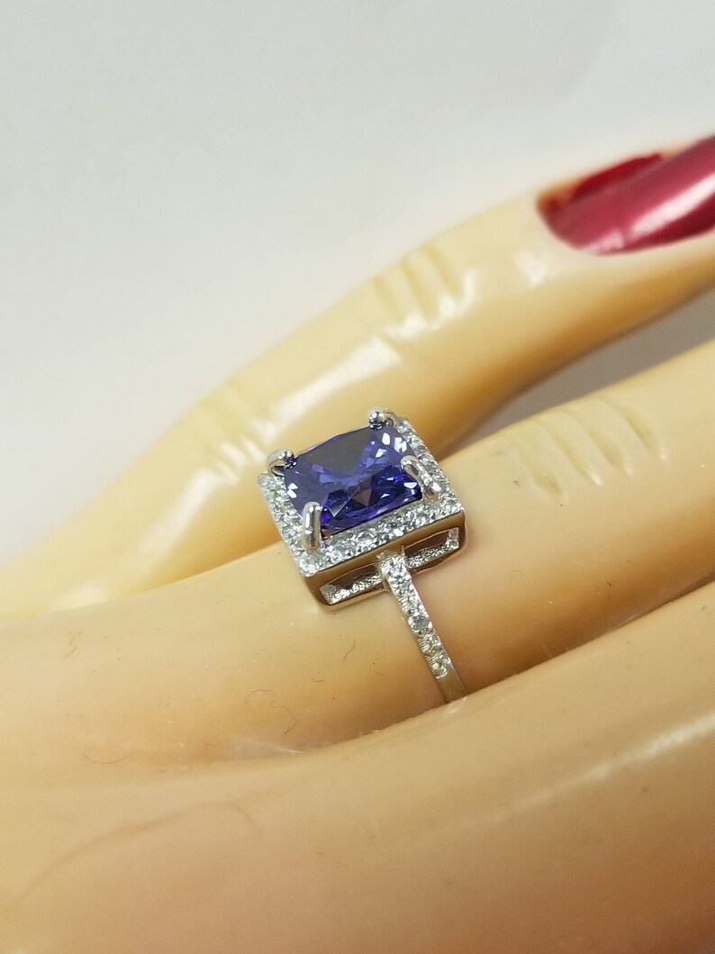 Op de afbeelding: Een zilveren ring met een grote, vierkante, paarse edelsteen omringd door kleinere, heldere edelstenen.