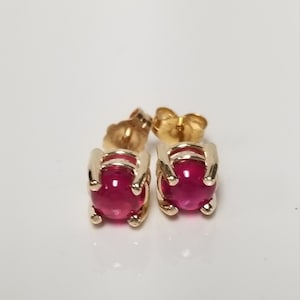 Op de afbeelding: Een paar goudkleurige oorstekers met levendige, ronde, rode edelstenen. De oorbellen hebben een klassiek ontwerp met een vier-klauwzetting. De oorbelruggen zijn ook goudkleurig.