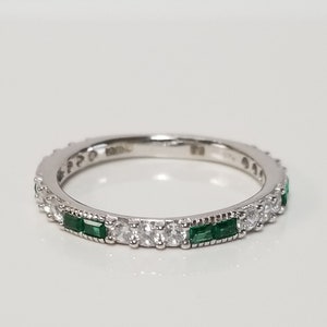 May include: A silver ring with alternating emerald-cut green and round white gemstones. The ring is set on a white background, showcasing the sparkle of the gemstones. The ring is a delicate piece of jewelry.